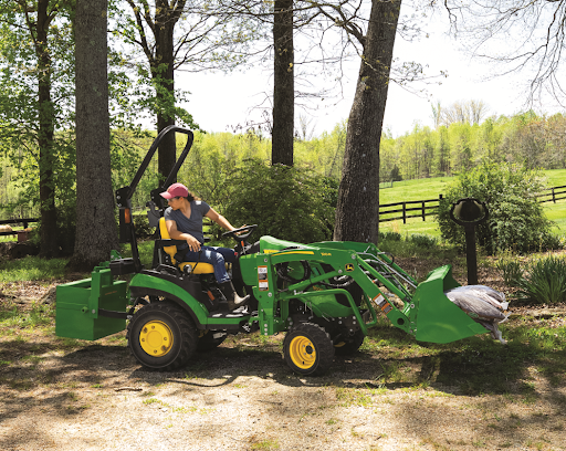 A woman uses a small tractor with a loader bucket to haul bags of garden soil 