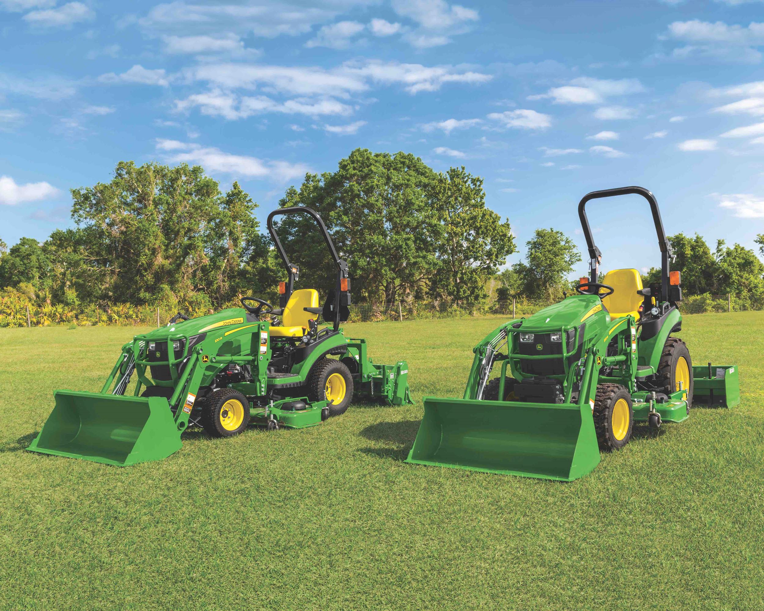 Two John Deere 1 Series tractors with front loaders parked on green grass with blue sky.