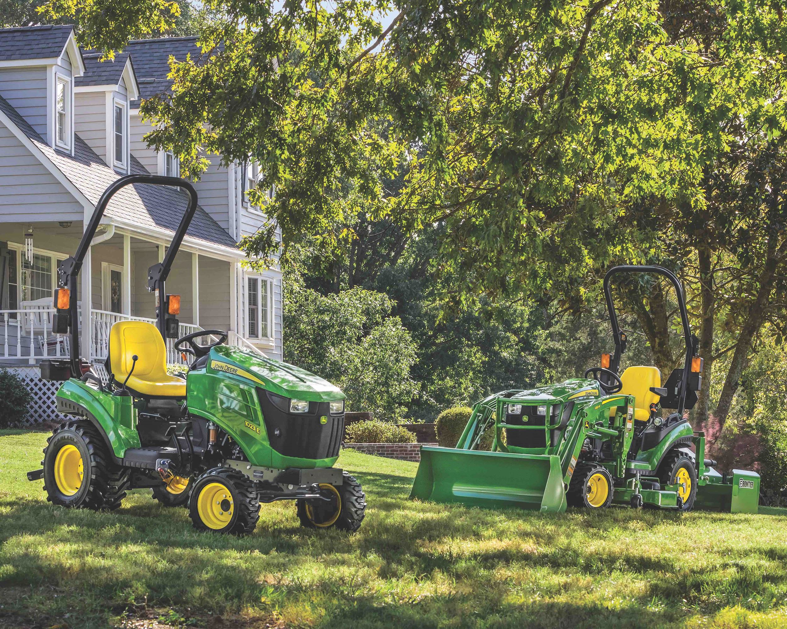 John Deere 1023E and 1025R sub-compact tractors are parked on a green lawn in front of a beautiful h