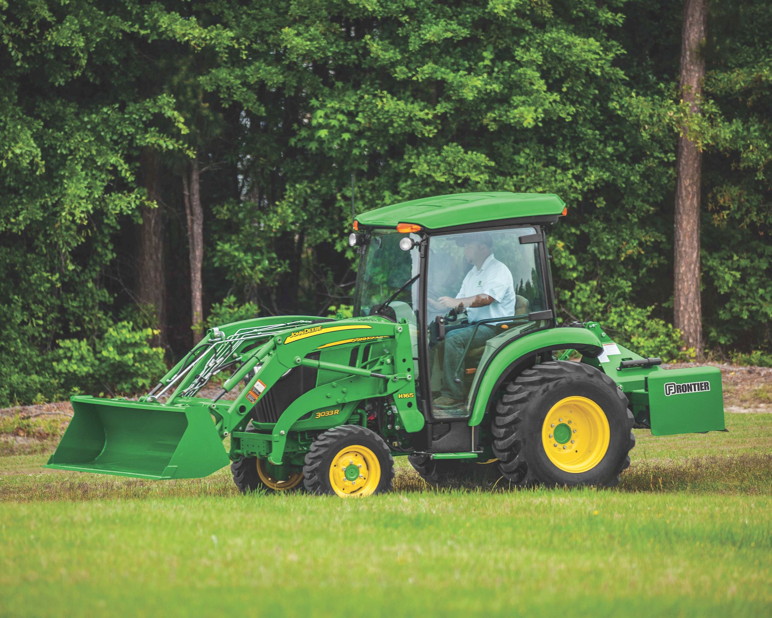 A John Deere 3033R compact utility tractor with a cab, H165 loader and box blade drives across green