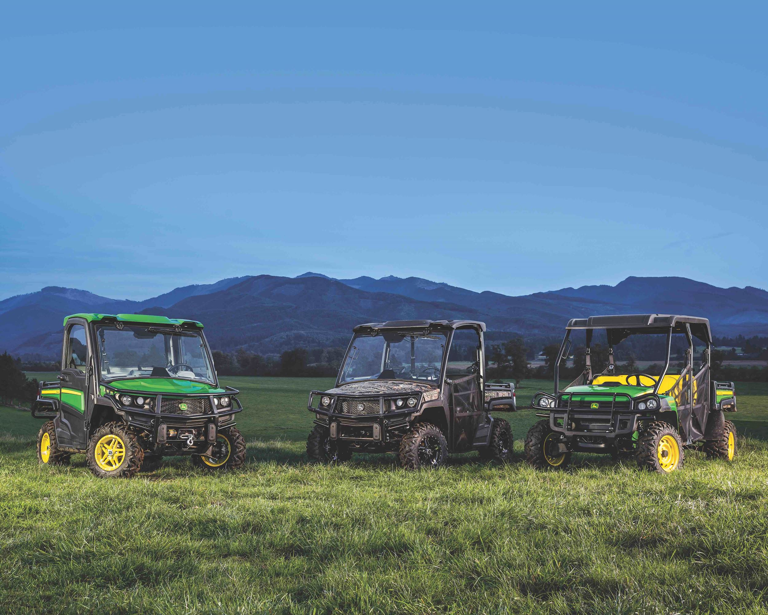 Three different John Deere Gators are parked on green grass with a mountain range in the background.