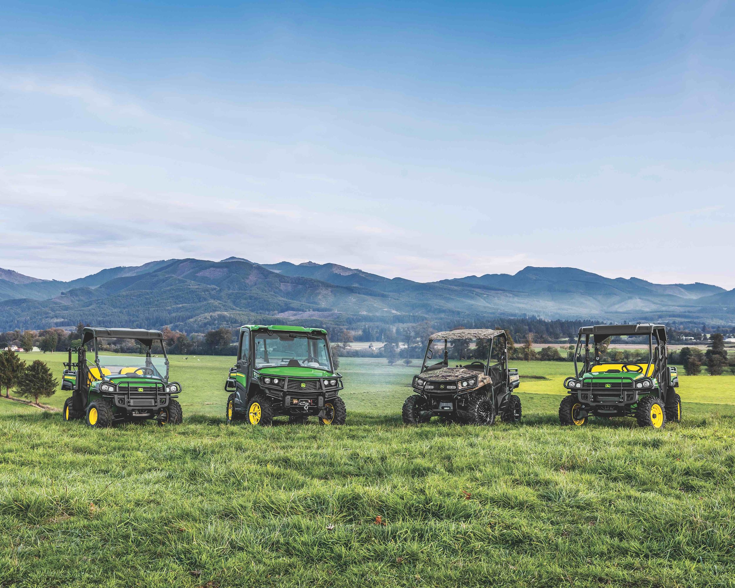 Four John Deere Gator utility vehicles are parked on green grass with a blue sky & a mountain range.