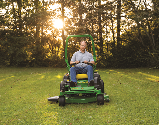 A man mows the grass using a John Deere zero turn mower. A row of trees is behind him and the sun is