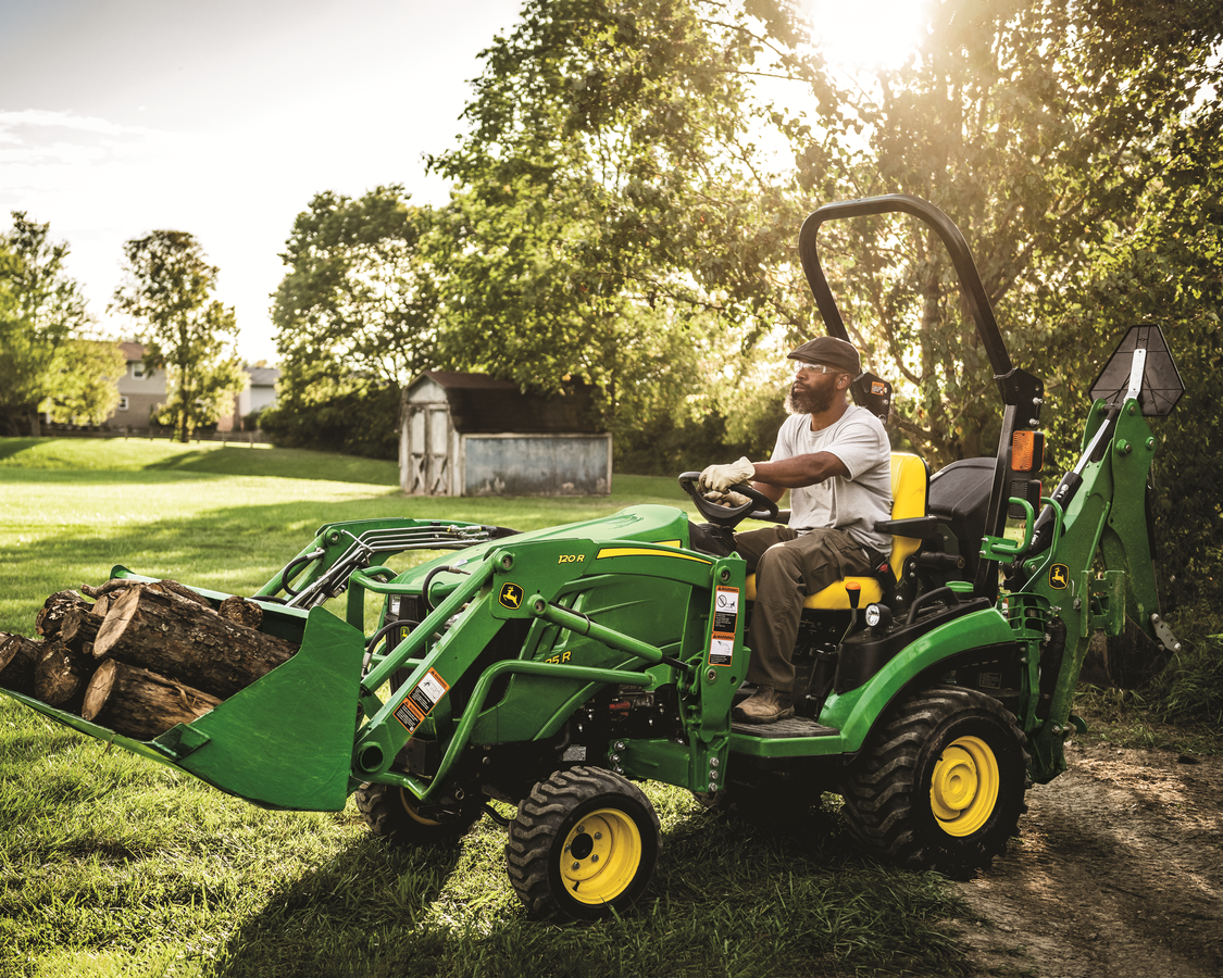 A bearded man in a cap, t-shirt, and gloves operates a green John Deere compact tractor