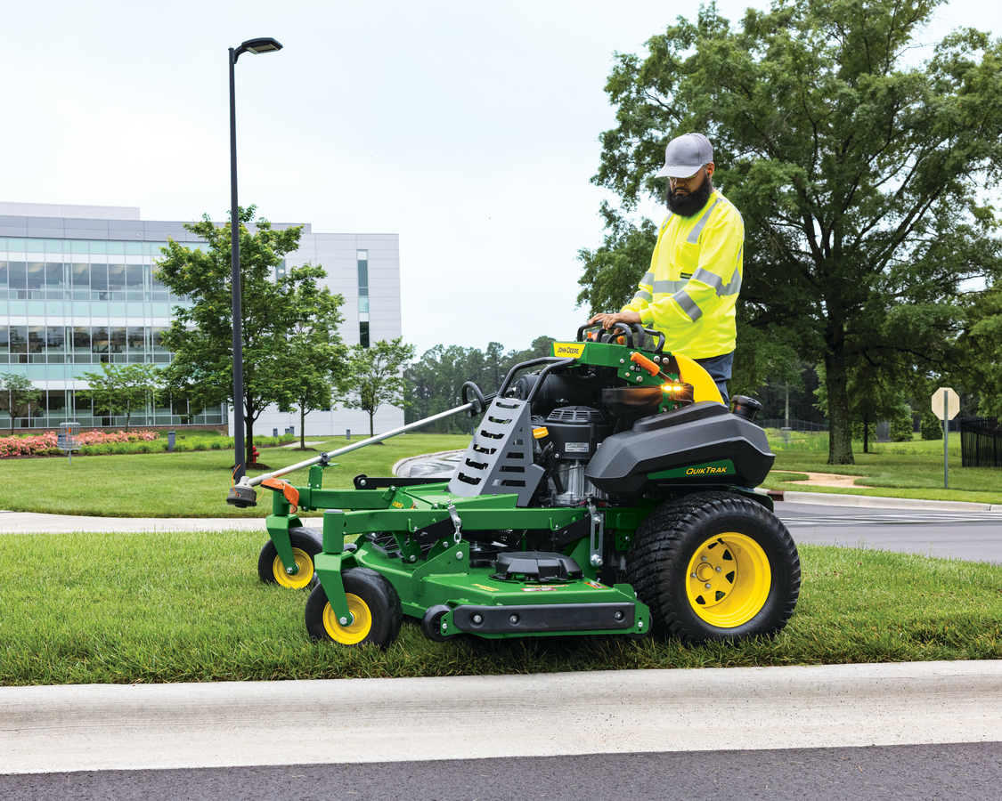 A person in a high-visibility yellow jacket and gray cap operates a green and black John Deere QuikT
